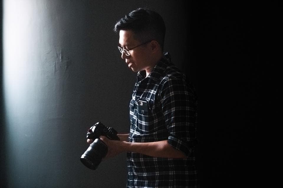 這張照片是一個戴眼鏡的男人在黑暗的房間裡手持相機,他穿著格紋襯衫,正在調整相機。