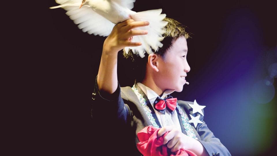 這張照片是一個年輕的魔術師,他正舉起一隻白鴿,準備表演魔術。他穿著西裝,戴著紅色領結,看起來非常專業。