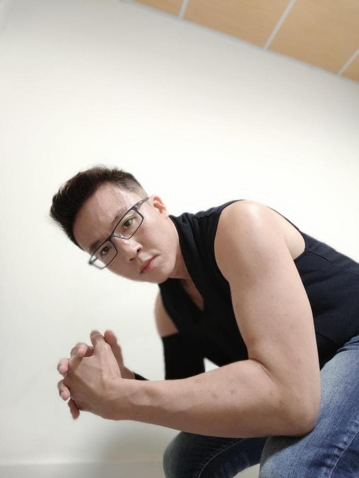 這張照片是一個戴著眼鏡的男人,他穿著黑色背心和牛仔褲,雙手放在膝蓋上,看起來很強壯。