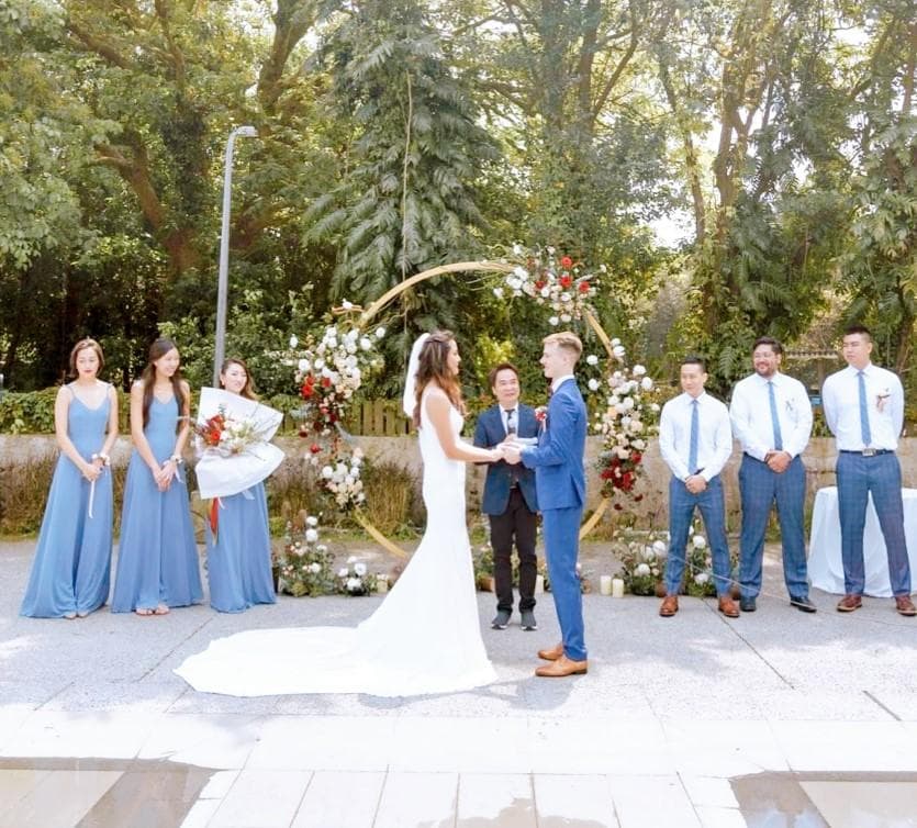 這張婚禮的照片是一對新人正在交換誓言,他們站在一個裝飾著鮮花的圓形拱門下,旁邊有幾位伴娘和賓客。