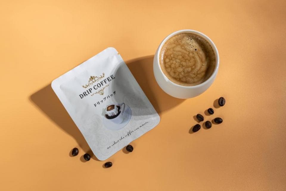 這張「滴濾咖啡」的照片是一個裝滿咖啡的白色杯子,旁邊有一個印有「DRIP COFFEE」的咖啡包裝,以及一些咖啡豆。