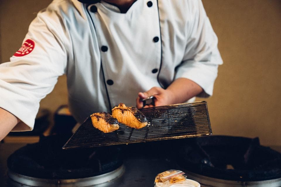 這張「廚師在烤魚」的照片是廚師正在烤兩片魚,他手裡拿著一個黑色的網子,上面放著烤好的魚。