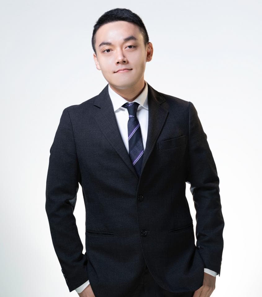 這張「穿西裝的年輕男子」的照片是一位穿著深色西裝、白色襯衫和紫色領帶的男子,他站在白色的背景前,表情自然。