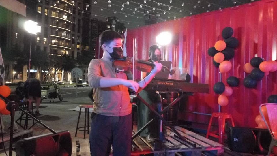 這張照片是一個年輕人正在演奏小提琴的畫面。他穿著深色衣服,戴著口罩,神情專注,看起來很有音樂天賦。他正在用右手拉弓,左手按弦,演奏出優美的旋律。背景是一面紅色的牆壁,牆上裝飾著一些氣球,營造出歡快輕鬆的氛圍。