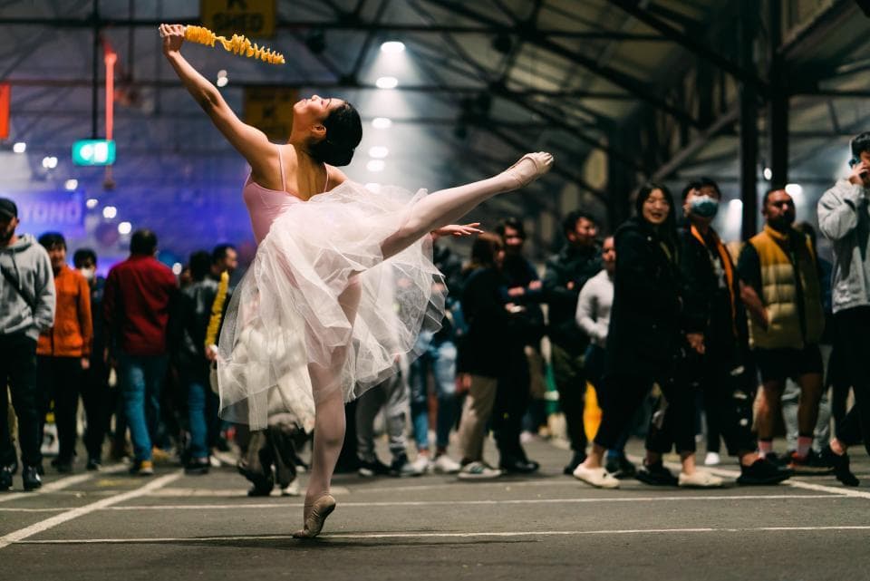 1) 照片中有一位女芭蕾舞者,身穿白色芭蕾舞裙,正舉起雙手並向後踢腿,在她面前有一群觀眾