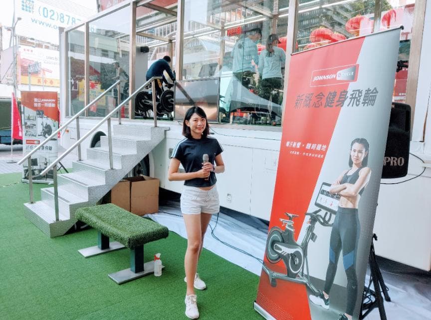 這張「健身器材活動」的照片是一個女性主持人站在一個白色貨車旁,她手持麥克風,正在介紹一輛室內自行車。