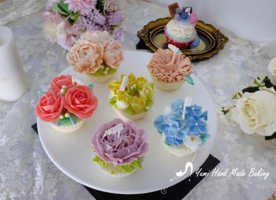 這張照片是六個以鮮花為主題的杯子蛋糕,每個杯子蛋糕都裝飾著精緻的花卉,從粉紅色玫瑰到藍色繡球花,每個花朵都栩栩如生,令人驚嘆。