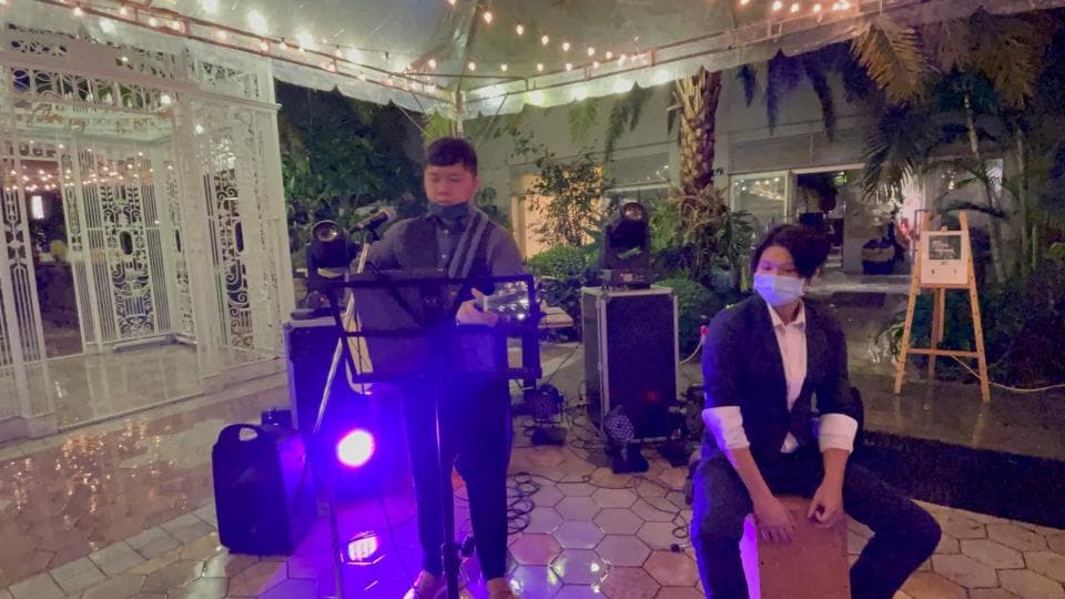 這張「戶外音樂表演」的照片是,兩個音樂家在一個戶外場地演奏,其中一位音樂家戴著口罩,背景是一棟白色建築和一些綠色的植物。