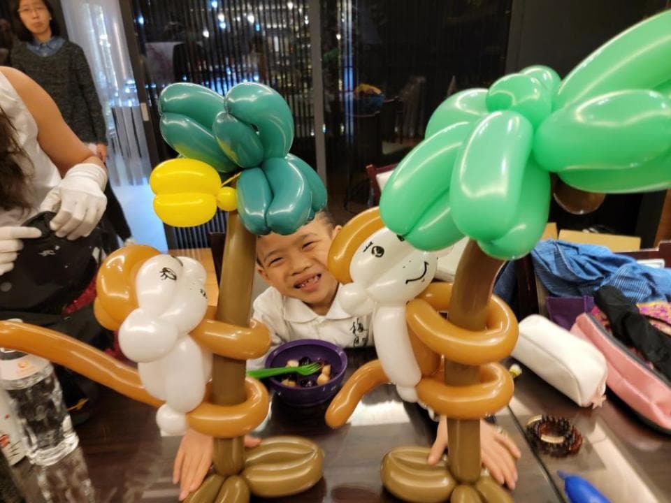 這張照片是一個小朋友坐在桌子旁,看著兩個氣球動物,一個是猴子,另一個是樹,他們玩得很開心。
