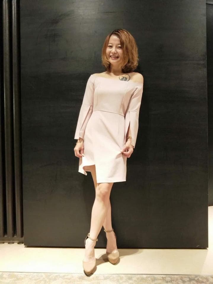 這張照片是一位穿著淺粉色連衣裙的年輕女子,她站在黑色牆壁前,面帶微笑,看起來很優雅。