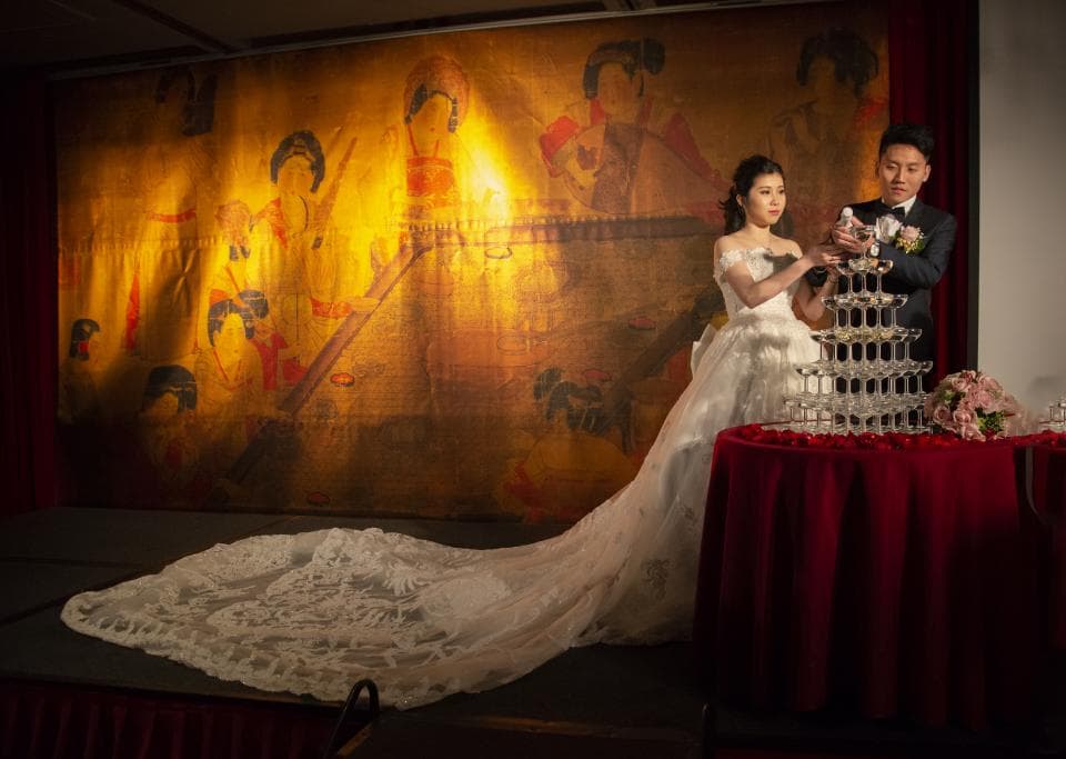 這張婚禮照片是一對新人正在一起開香檳塔,背景是一幅金色調的壁畫,象徵著幸福和喜慶。