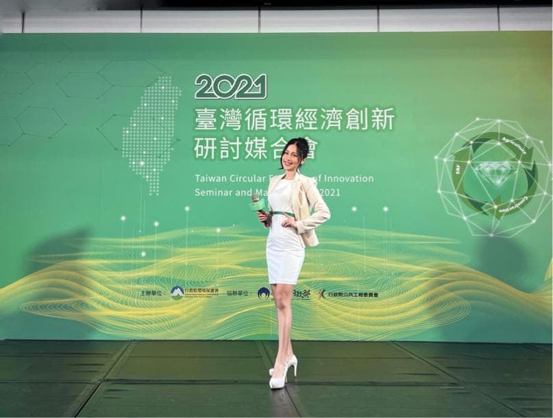 這張「臺灣循環經濟研討會」的照片是一名女性站在舞臺上,她穿著白色洋裝,正在微笑,背景是一塊綠色的布幕,上面寫著「2021 臺灣循環經濟研討會」。