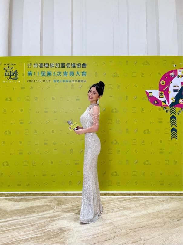 這張「頒獎典禮」的照片是一個穿著閃亮禮服的女子,她手拿獎盃,站在綠色的背景板前,背景板上有許多文字和圖案。