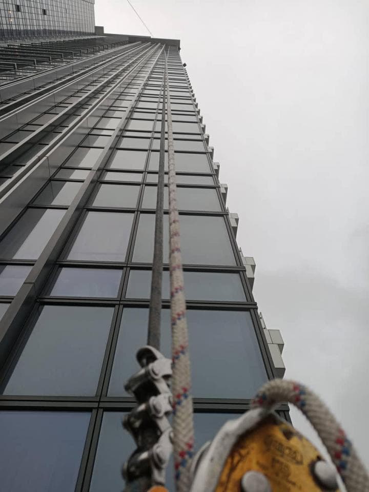 這張照片是一棟高聳的摩天大樓,畫面中有一條粗大的繩索從頂端垂下,繩索上連接了一個金屬滑輪,讓人聯想到建築工人正在進行高空作業。