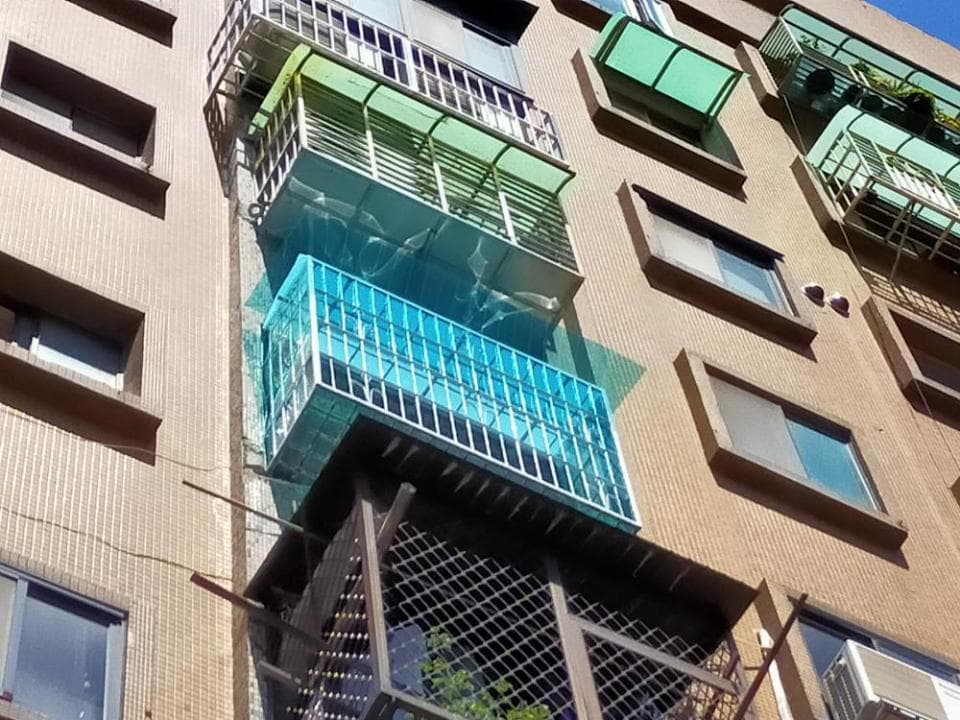 這張「建築物外牆」的照片是一個有許多窗戶的建築物,其中有一個陽臺被藍色的鐵欄杆圍起來,陽臺上還有一個綠色的遮陽棚。