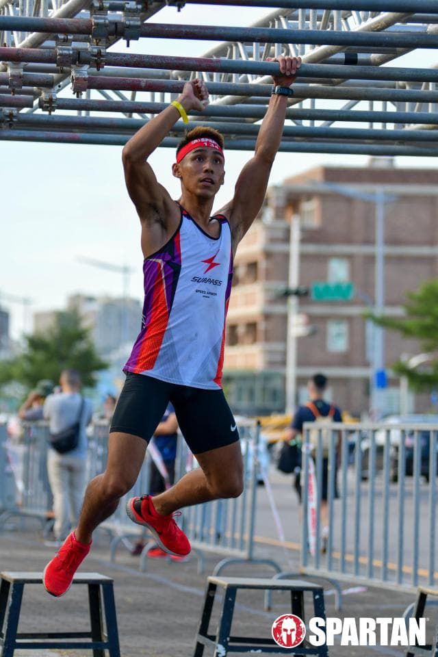 這張照片是一個年輕男子在戶外障礙賽中奮力穿越一組金屬杆。他雙手抓住杆子,雙腳離地,身體懸在空中,展現出驚人的力量和敏捷性。他的表情堅定,眼神充滿決心,展現出積極向上的精神。