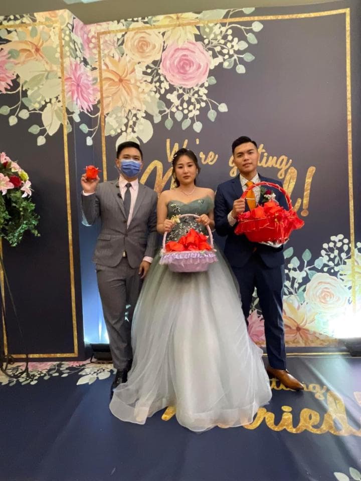 這張「婚禮」的照片是三個人站在花牆前,中間穿著白紗的女子手拿花籃,左右兩邊是新郎和伴郎,他們都拿著禮物。