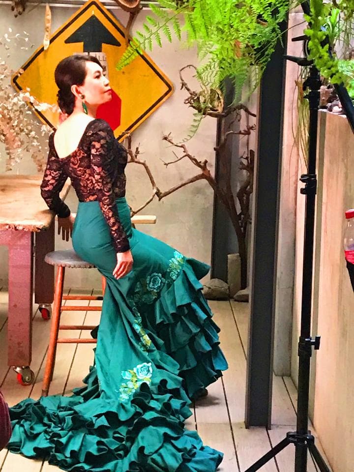 這張照片是一個穿著翠綠色荷葉邊長裙的女子,她站在一個室內空間,背景是一個木頭桌子和一個木頭凳子。