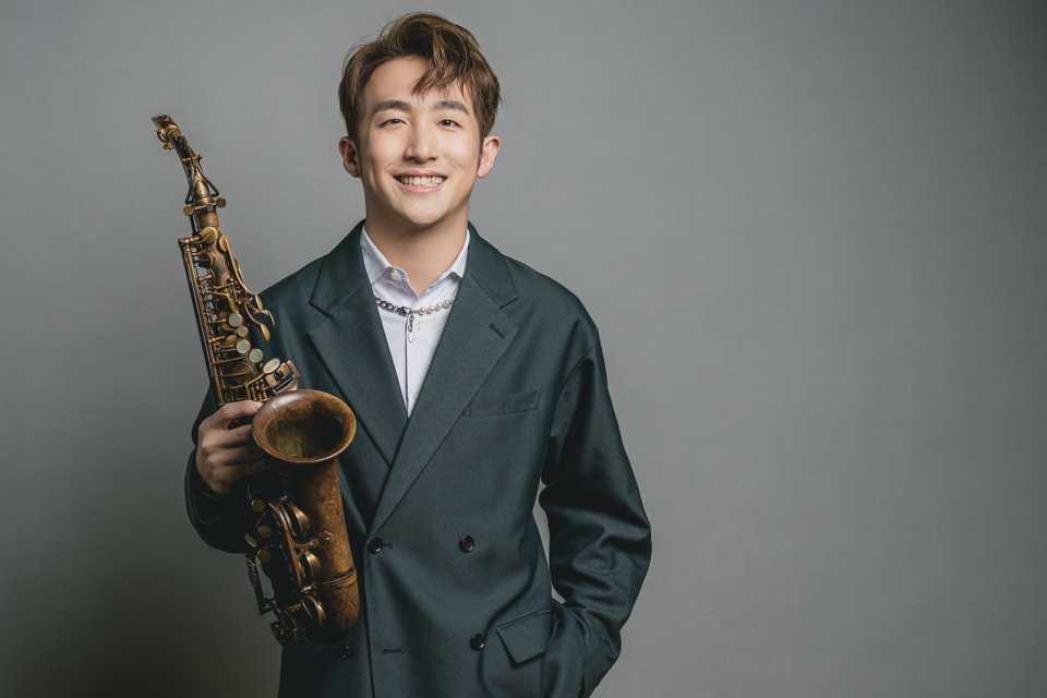 這張照片是一個穿著西裝的年輕男子,他拿著一把薩克斯風,正對著鏡頭微笑。