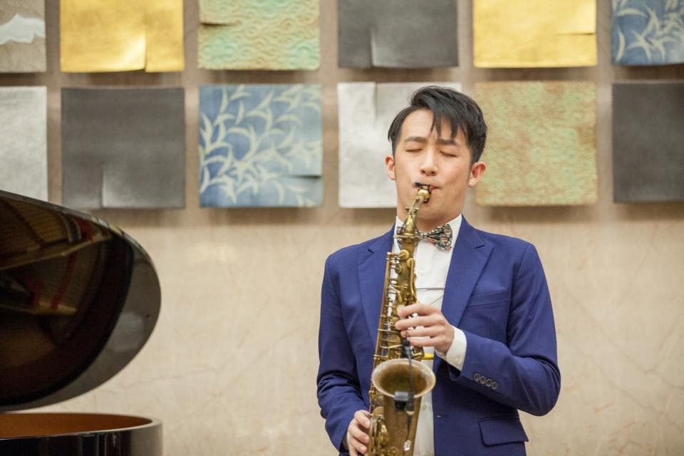 這張照片是一個年輕男子吹奏薩克斯風的畫面。他穿著深藍色的西裝,並打著領結,專注地吹奏著薩克斯風,背景是一面裝飾著瓷磚的牆壁。