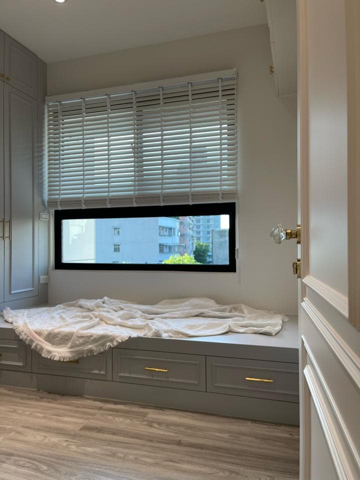 1) 照片展示了一間房間的窗戶和床鋪,床鋪上有一床白色床單,窗戶上是白色百葉窗,窗外可以看到高樓建築,窗戶下方是一個木質平台,平台上有一個抽屜櫃