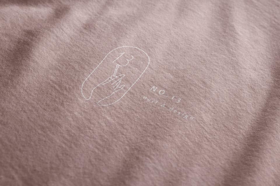 這張照片是一件印有白色圖案的淡紫色T恤。圖案是一個橢圓形,裡面有一隻手拿著一根火柴。橢圓形下方寫著「NO.13」,可能是衣服的編號或系列名稱。