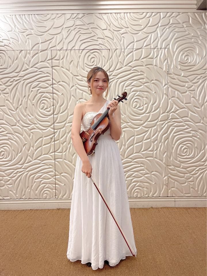 這張照片是一個穿著白色禮服的年輕女子,她正在演奏小提琴。她看起來很優雅,背景是玫瑰花牆,為照片增添了浪漫的氛圍。