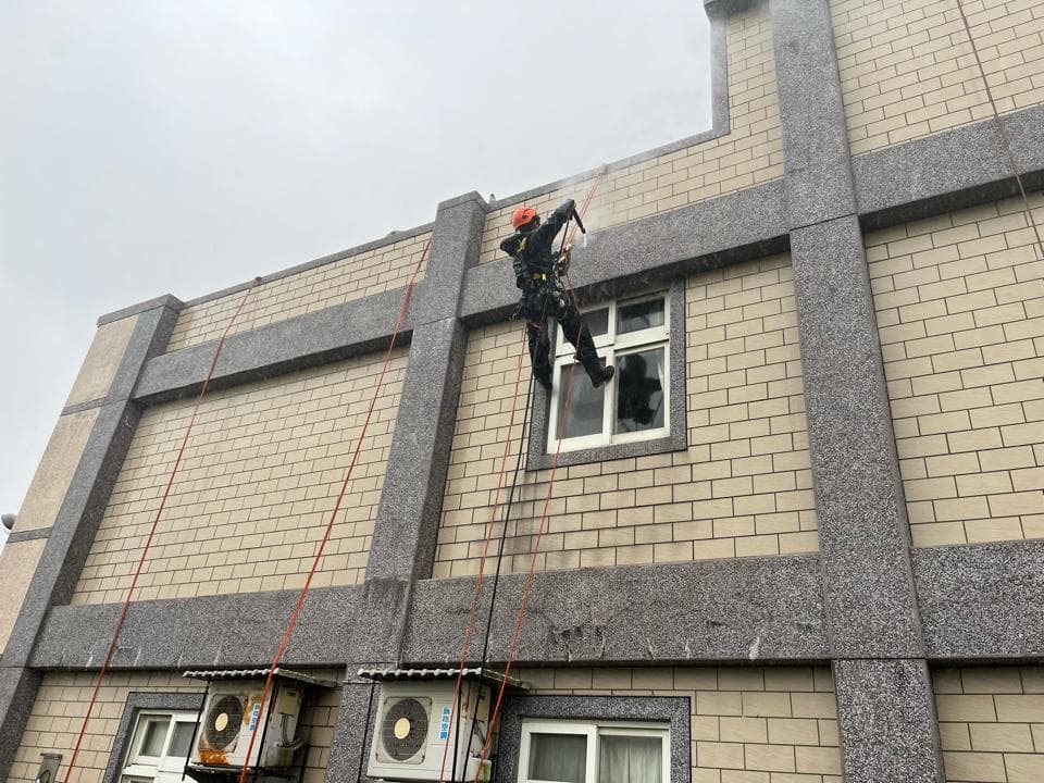 1) 照片顯示一名身穿黑色制服,戴著安全帽的工作人員,正懸掛在建築物外牆上,使用繩索下降,進行外牆清潔工作