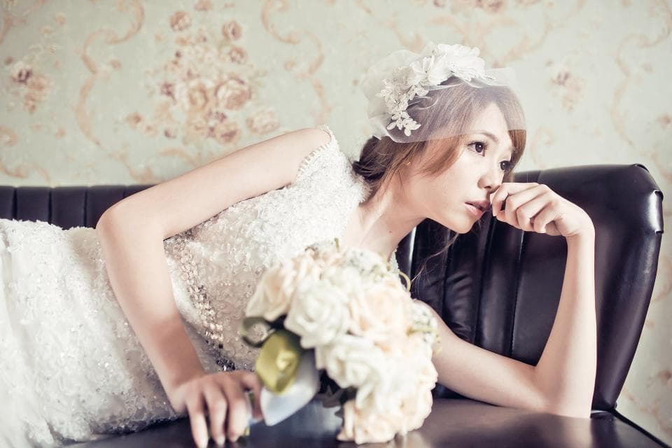 這張照片是一個穿著婚紗的新娘,她坐在沙發上,手裡拿著一束白色玫瑰花,看起來很幸福。