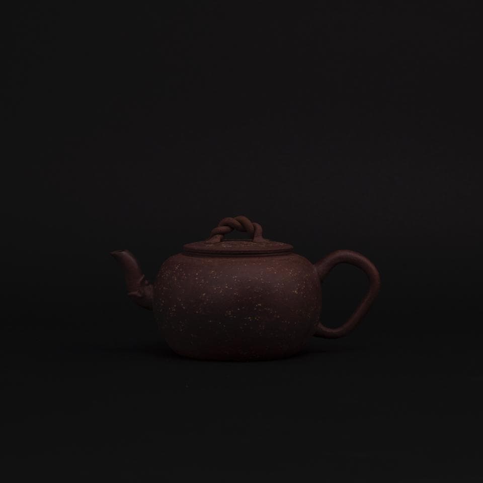 這張照片是一個紫砂壺,壺身呈現深紅色,壺嘴和壺把均為彎曲設計,壺蓋上有一個繩狀的提手。壺表面佈滿了細小的點狀紋理,增添了古樸雅緻的質感。