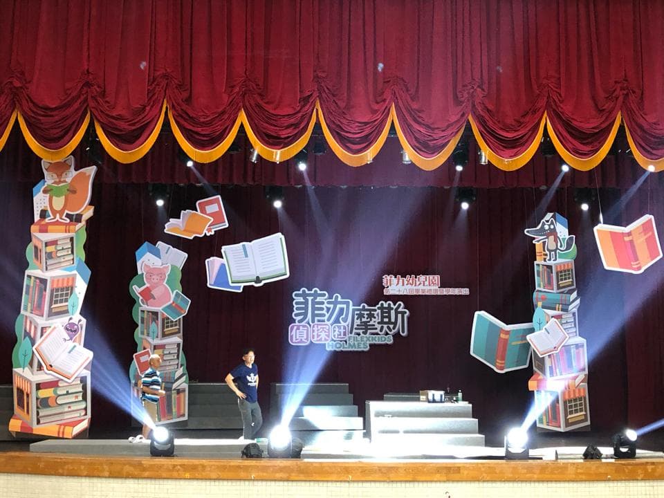 照片展示了一個舞台,舞台上佈置著童書的圖案,舞台中央有兩個穿著休閒服的成人,舞台前方擺放著一個小型講台