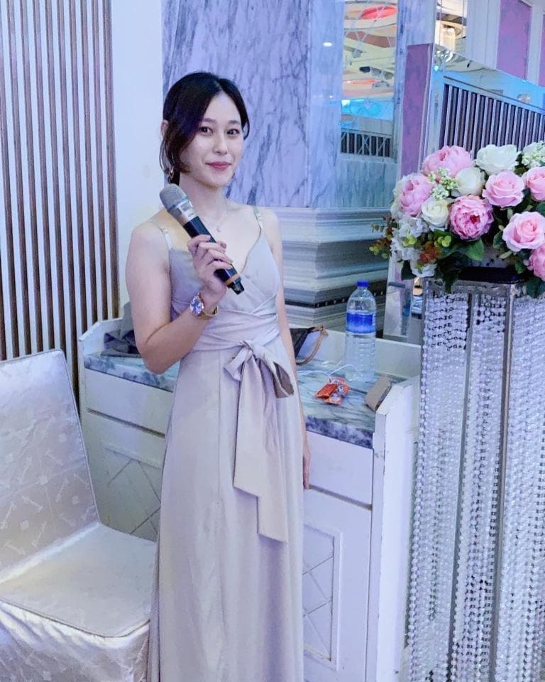 這張「婚禮」的照片是一個穿著淺灰色禮服的女人,她拿著麥克風,站在白色桌子的旁邊,旁邊有花束。