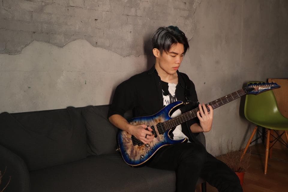 這張照片是一個年輕人坐在沙發上彈奏電吉他的照片。他專注地看著琴弦,並用手指撥動琴弦,彈奏出美妙的音樂。他身穿黑色外套,頭戴藍色帽子,看起來很有音樂才華。