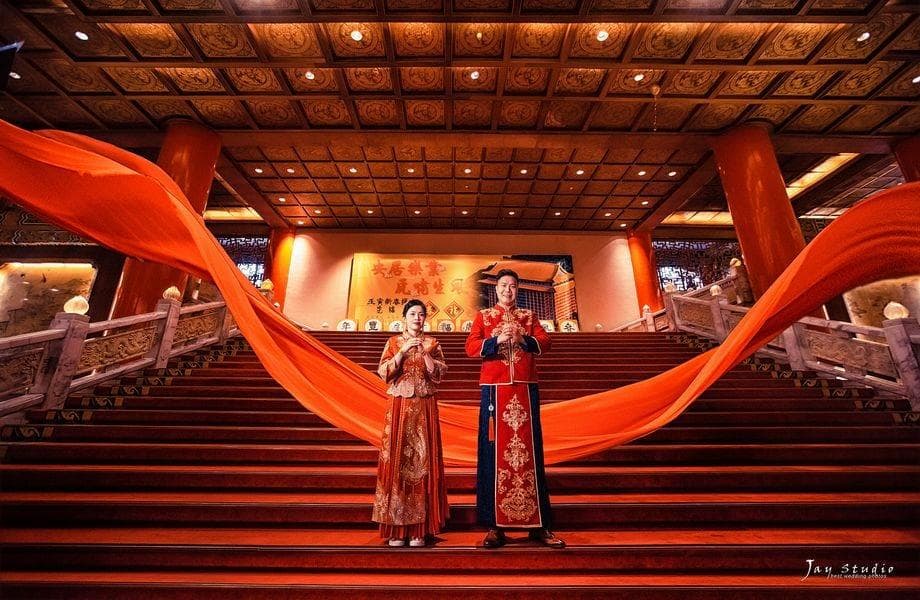 這張照片是一對身穿傳統服飾的男女,站在紅色絲綢佈景上,背景是華麗的宮殿式建築。