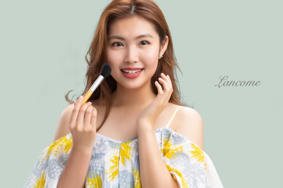 這張「蘭蔻」的照片是一個女人用化妝刷塗抹臉頰,她穿著淡黃色的衣服,背景是淡綠色的牆壁。