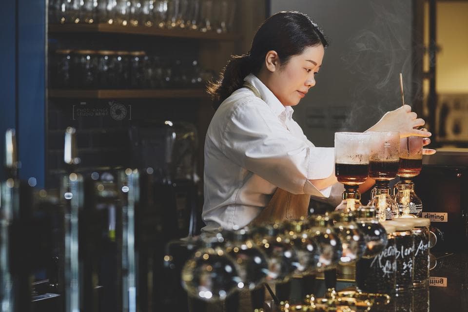 這張「咖啡廳」的照片是一個年輕女子正在調製一杯咖啡,她身穿白襯衫,專注地看著手中的咖啡杯。