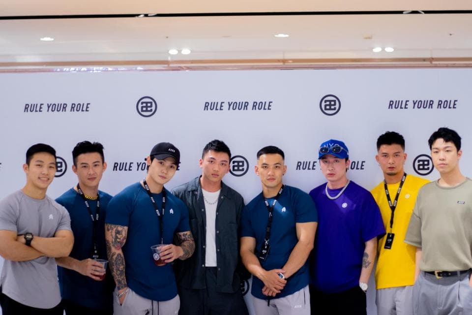 這張「一群男性站在牆壁前」的照片是七個男人穿著不同顏色的衣服,他們都看著鏡頭微笑。牆壁上寫著「RULE YOUR ROLE」和一些符號。