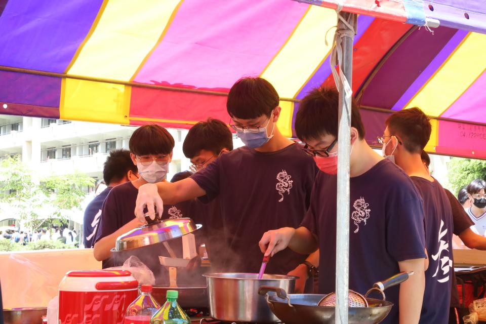 這張「一群人在戶外攤位上煮東西」的照片是,畫面裡有許多人正在煮東西,其中一個男生戴著口罩,穿著藍色衣服,正在攪拌鍋裡的東西。