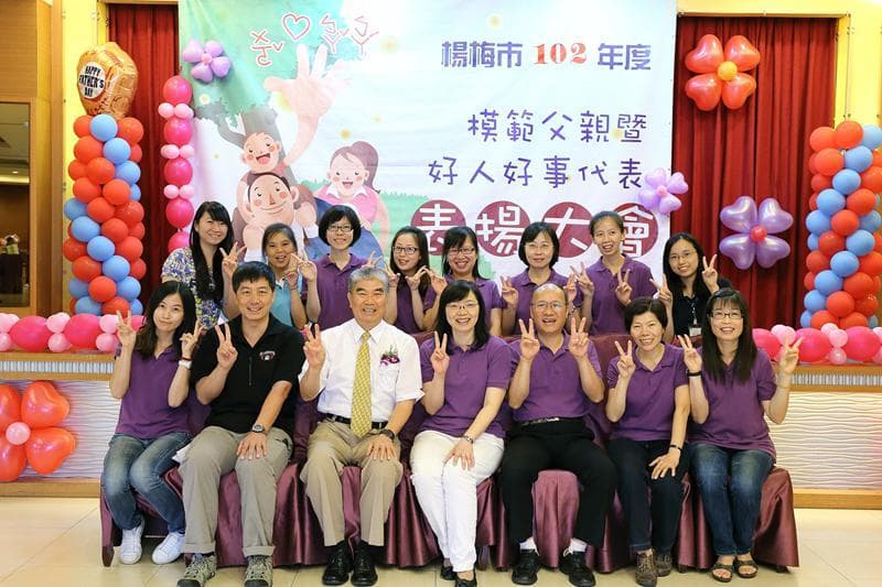 這張「一群人站在背景布前微笑拍照」的照片是,照片中的人們都穿著紫色的衣服,背景布上寫著「幸福 ‧ 親 親 ‧ 親善,好人好事代表」的標語。