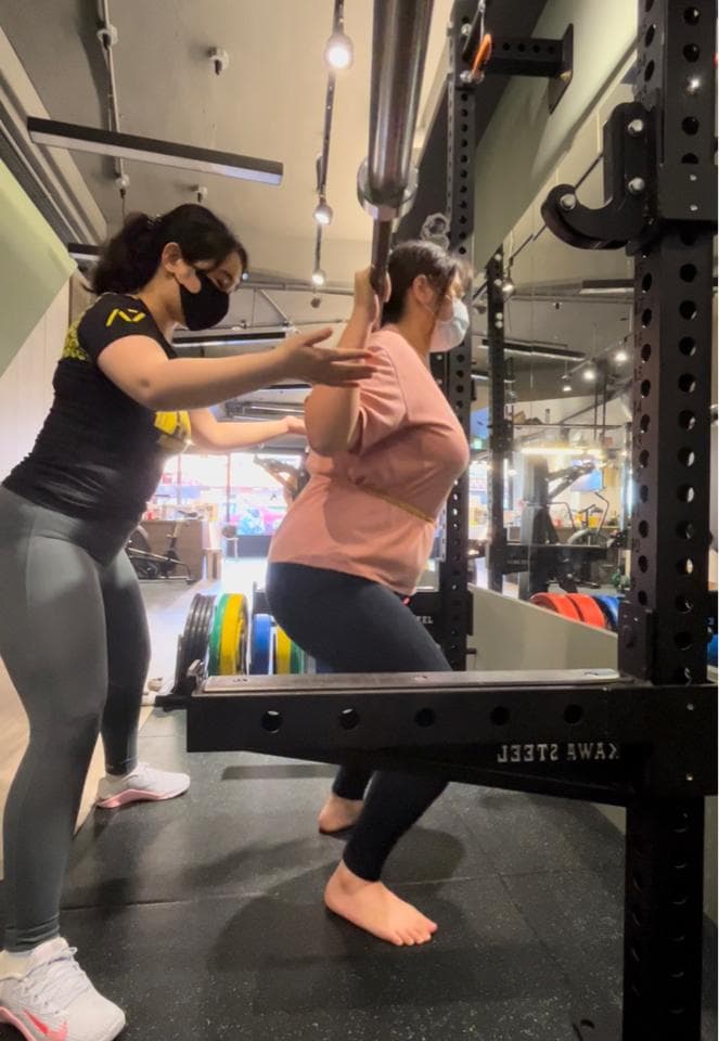 這張照片是一個健身房,照片中,一位女性正在進行深蹲訓練。 她正由另一位女性教練指導。 教練正觀察著她的動作,並給予她指導。 畫面中,兩人專注於訓練,展現了健身房中積極向上和努力不懈的精神。