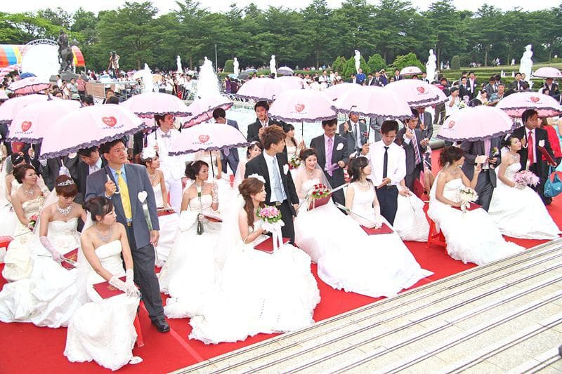 這張婚禮照片是許多穿著白色婚紗的新娘,她們站在紅色的地毯上,拿著捧花,旁邊有許多穿著西裝的男士。