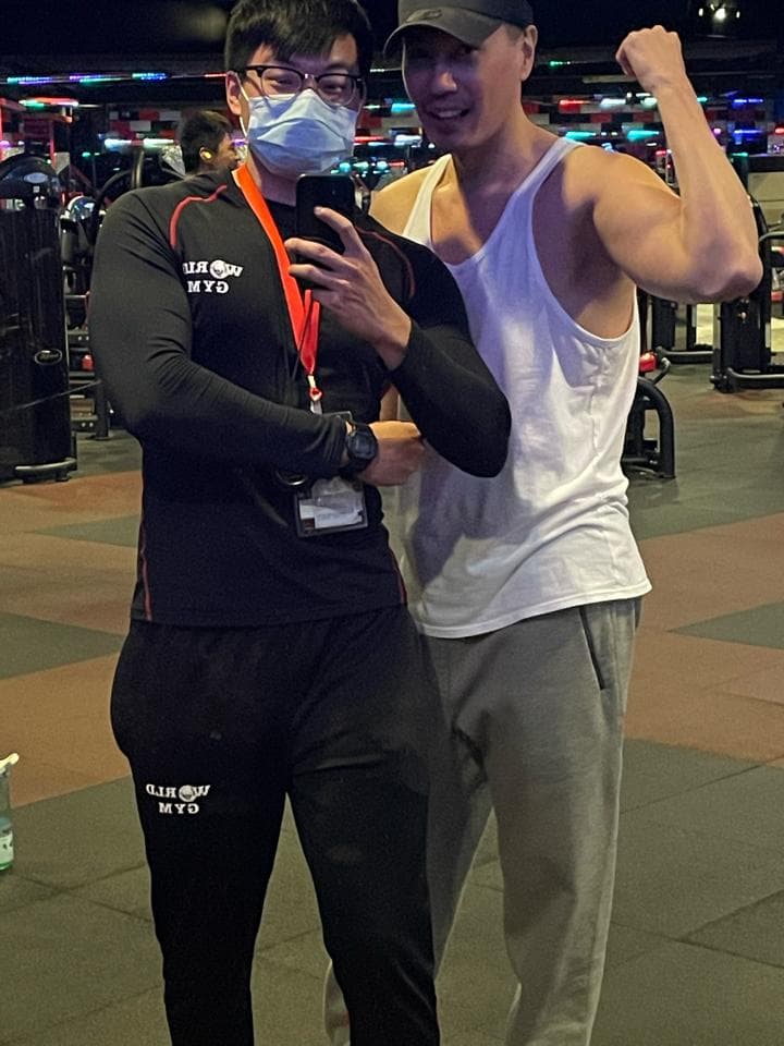 這張「健身房」的照片是兩個男生,他們在健身房裡。左邊的男生穿著黑色運動服,戴著口罩,右手拿著手機,右邊的男生穿著白色背心和灰色運動褲,右手舉起來。