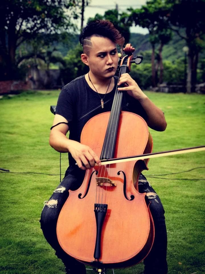 這張照片是一位年輕男子坐在草地上,手持大提琴。他穿著黑色的T恤,正專注地演奏。他的表情專注,手勢流暢。大提琴的琴身是木質的,有著光滑的表面,琴絃被拉得筆直。
