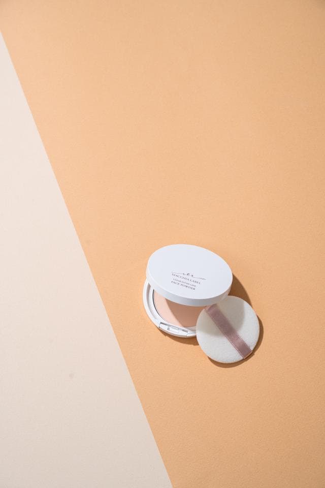 這張照片是一盒粉餅,粉餅盒是白色的,上面印有黑色的字。盒子旁邊有一個白色粉撲,粉撲上面有一條粉紅色的線。