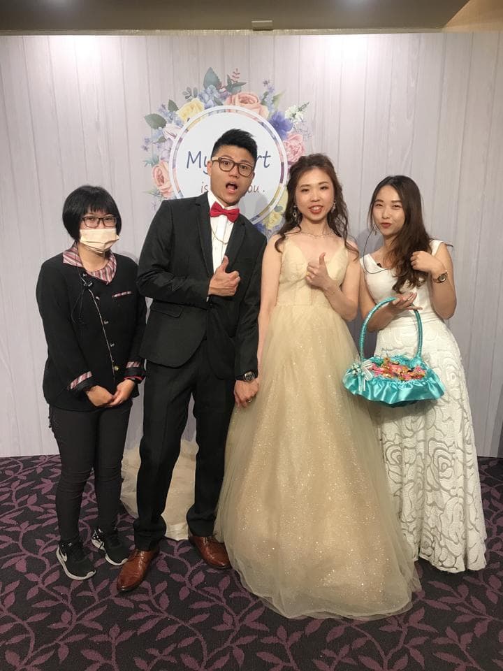 這張「婚禮」的照片是三個人站在花牆前,中間穿著婚紗的女孩手拿著一個藍色的花籃,右邊穿著白色洋裝的女孩,左邊穿著西裝的男孩。