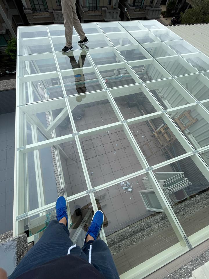 這張「玻璃屋頂」的照片是一個用玻璃和金屬框架製成的屋頂,看起來很堅固。屋頂下有灰色磚塊,一個人的腳和鞋子出現在玻璃上。