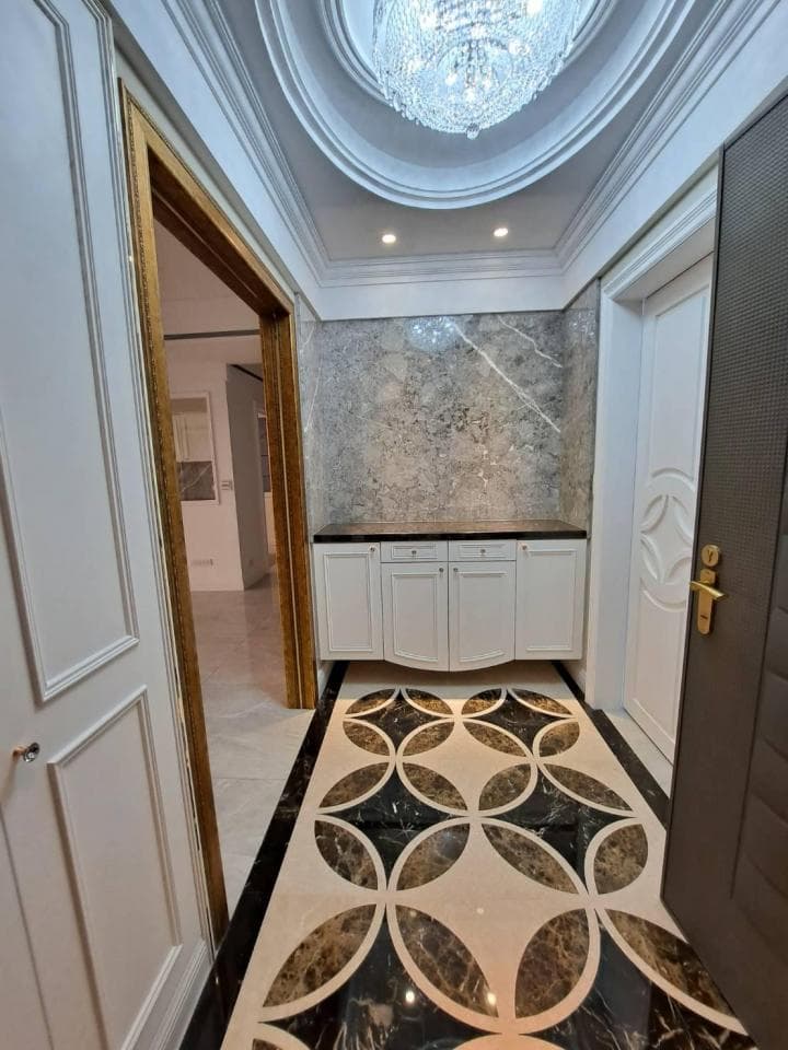 1) 這是一張室內設計照片,照片中可以看到一個有著精緻裝潢的玄關,地板是白色大理石拼花,牆面是灰色大理石,玄關上方有一個水晶燈,玄關左側有一個白色木門,右側有一個木門,中間是白色櫥櫃