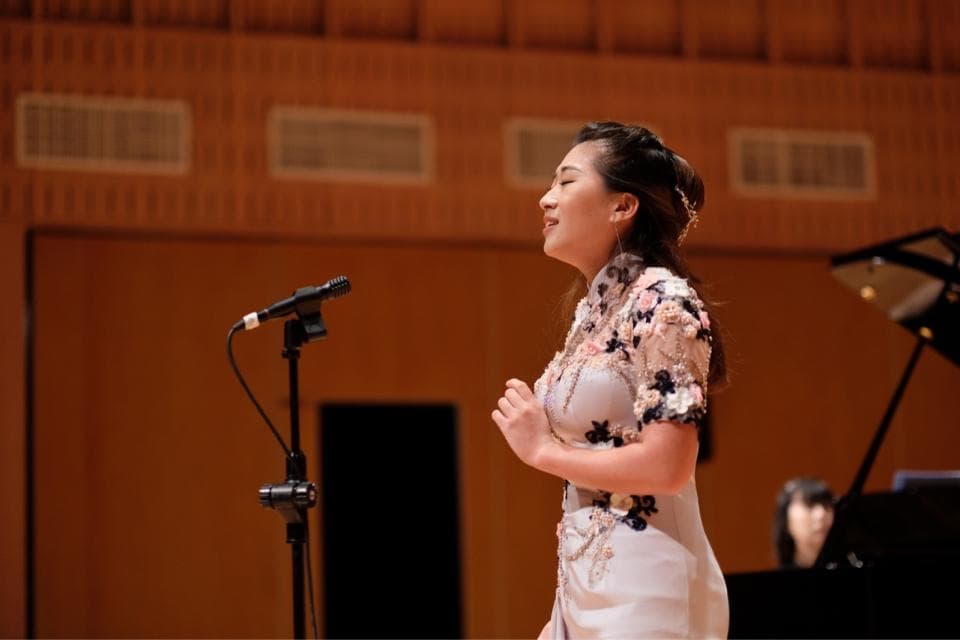 這張照片是一位穿著淡粉色花朵圖案連衣裙的年輕女性,正在演唱,她面前有一個麥克風。