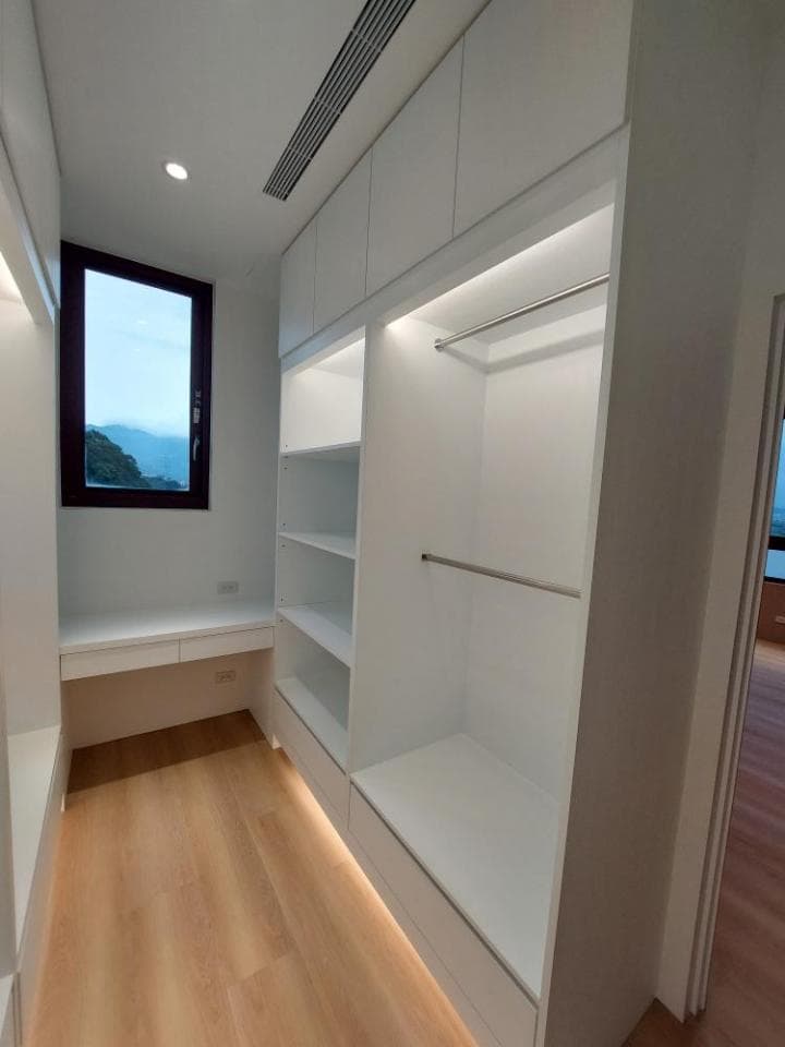 這張照片是一個現代化的衣帽間,裡面的牆壁和櫥櫃都是白色的,地面鋪著淺色的木地板,有一個大窗戶,可以讓陽光照射進來。最顯眼的是衣帽間的燈光設計,使用了隱藏式的燈條,讓衣帽間顯得更加明亮和高級。