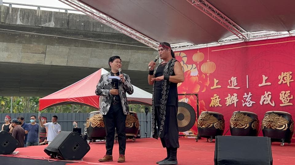 這張照片是兩個男人站在舞臺上講話,他們身後有大型鼓,臺下還有觀眾,舞臺上掛著紅色布條,上面寫著「上道 | 上輝 誠摯送祝福」,畫面充滿喜慶的氣氛。